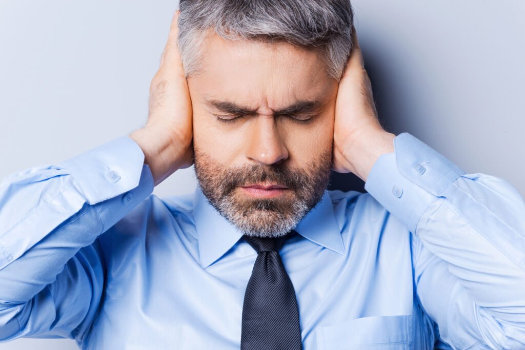 Feeling stressed and overworked. Frustrated mature man in shirt and tie covering ears with hands and keeping eyes closed while standing against grey background